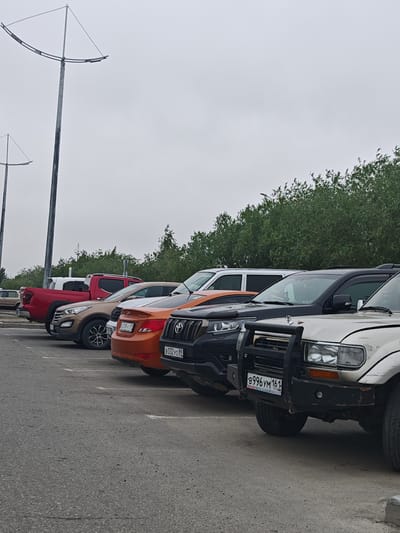 A row of parked vehicles, including several SUVs and a pickup truck, are visible in an outdoor parking lot. The sky is overcast, and a line of green trees forms the background. Two tall, industrial-looking poles with a triangular structure at the top are present on the left side of the image. License plates are visible on some of the vehicles, and one license plate displays "B996YM 161 RUS," indicating it is registered in Russia. The ground is paved asphalt. The scene appears to be a typical parking area in Novy Urengoy, Russia, given the presence of Russian license plates and the general environment. There are no people visible in the image.