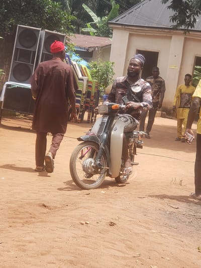 The image captures a scene in Umundugba, Nigeria, featuring a man seated on a motorcycle. He wears a patterned shirt, a cap, and appears to be looking directly at the camera. A second individual, dressed in a brown robe and wearing a red cap, stands nearby. In the background, there are other people, some dressed in yellow, near a building. A stack of folded chairs is visible, along with large speakers. The ground is dirt and sand.