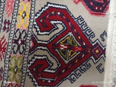 The image shows a close-up, overhead view of a section of a rug with a beige background and intricate geometric patterns in red, black, white, yellow, and green. The rug has a fringe on the right side. A timestamp "01.07.2026 01:44" and the text "REDMI 15C" are visible in the lower corners. The rug is likely located indoors, possibly in a home or shop in Balkanabat City, Turkmenistan, as indicated by the text. The lighting suggests it is daytime, but the exact environmental context is limited by the close-up nature of the shot.