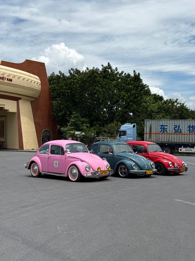 The image shows a line of four vintage Volkswagen Beetles parked on a paved surface, likely a street or parking lot. From front to back, the cars are pink, teal, blue, and red. The pink car has a prominent sticker on its side with a circular design. The teal and blue cars each have yellow license plates. The red car has a license plate with red text. Behind the cars, a large white shipping container with Chinese characters ("东弘物") is visible, along with a blue truck cab. A tall green tree line runs along the back of the vehicles, and power lines are visible against a cloudy blue sky. To the left of the cars, a building with a brick facade and a cream-colored arched section with decorative molding is partially visible. Text on the building reads "VIỆT NAM GATE". The overall scene suggests a bright, daytime setting. The cars are the central focus, arranged in a neat row, implying they are on display or part of a collection. The presence of the shipping container and truck suggests a commercial or industrial area, possibly near a border crossing, given the "Lao Bảo Commune, Vietnam" context. The cars' vibrant colors and classic design evoke a sense of nostalgia and style.