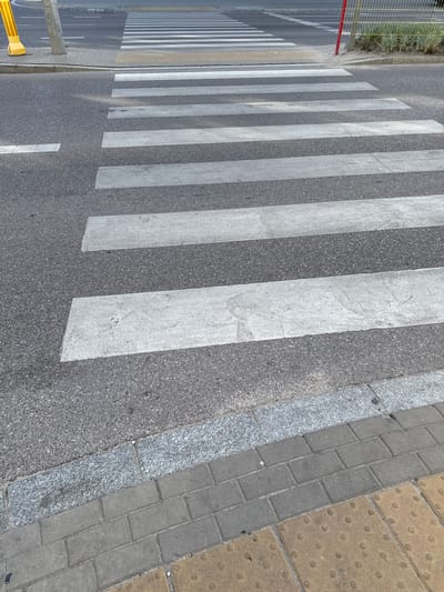 The image shows a pedestrian crossing in Białystok, Poland. The crossing is marked with white stripes on asphalt. The sidewalk is paved with cobblestones, some of which have tactile paving for the visually impaired. A yellow bollard with reflective stripes is visible on the left side of the image, near the curb. In the background, a metal fence and some vegetation can be seen. The scene appears to be a typical urban street environment.
