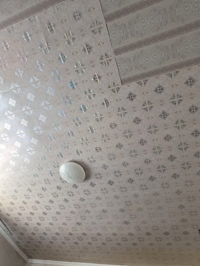 This media file shows an indoor ceiling with patterned wallpaper and a light fixture. The wallpaper has a repeating geometric design, with some sections featuring a more intricate, ornamental pattern. The colors are muted, with shades of grey and beige dominating. A circular, white light fixture with a ribbed texture is attached to the ceiling. The overall impression is of a typical interior space, possibly a room in a residential or commercial building. There is no indication of the time of day or weather outside. The location is identified as Қанабек, Kazakhstan. There is no visible text in the image.