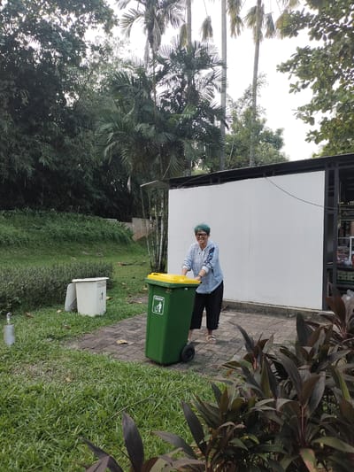 An individual with short, teal-colored hair and glasses is depicted smiling while pushing a green wheeled garbage bin with a yellow lid on a paved brick pathway in Gunung Putri, Indonesia. The person wears a light blue and white patterned long-sleeved shirt, black pants, and white sandals. The green bin features a white rectangular sticker depicting a person discarding trash into a bin, with accompanying text "KRIBOW." To the left, a white open-top bin labeled "CITRA GRIA" with the number "1" rests on a patch of grass. A white wall with black metal framing, indicative of an outdoor structure or stall, occupies the midground-right. The surrounding environment is characterized by dense green vegetation, including tall palm trees, various broad-leaved trees, and bamboo-like plants in the background. Lush green grass covers the ground, with darker reddish-purple foliage visible in the immediate foreground-right. The scene is illuminated by bright daylight.