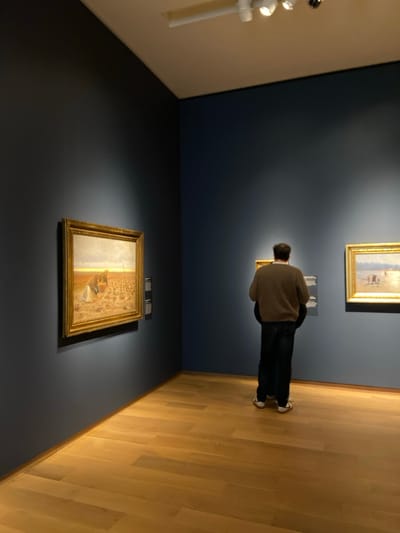 The image shows a man standing in a gallery room. He is wearing a brown sweater and blue jeans, looking at a painting that is hanging on the wall to his right. The painting is a landscape scene and has a golden frame. The man has a woman standing to his left who is partially obscured. The gallery has blue walls, a white ceiling with recessed lighting, and a wood floor. There are two other paintings hanging on the wall opposite the man. The gallery appears to be located in Greenwich, Connecticut, United States. The room is dimly lit. The man is the only person visible in the image. The environment is serene and quiet. The lighting is soft and even. The overall impression is one of contemplation and quiet appreciation for art.