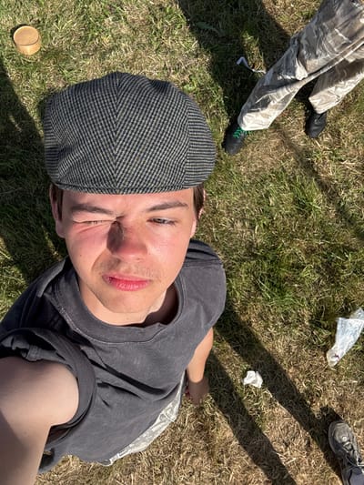 A person wearing a gray newsboy cap and a faded gray t-shirt lies on the grass, gazing up at the camera. Their head is tilted slightly. Sunlight illuminates their face, accentuating their features. The grass around them is a mix of green and brown. Parts of other people's legs and feet appear near the top of the frame. A small, round object sits to the lower left of the person in the center. The overall scene has a casual, outdoorsy feel.