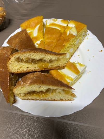 A plate of sliced cakes is placed on a dark brown patterned surface. The cakes appear to be a sweet pastry with a golden-brown crust and a light-colored interior. Some slices are filled with a translucent yellow jam or preserve. Another cake on the plate is yellow and appears to have white chunks or pieces mixed within it, and is also topped with white frosting and streaks of yellow. The plate is white and has an irregular, wavy edge. In the upper left corner, partially visible, is a clear plastic container with more baked goods inside. The lighting suggests an indoor setting. The scene does not provide any specific information that would allow for the localization of the city of Izberbash, Russia.