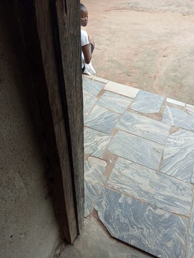The image captures a young woman sitting outside, appearing to be in contemplation. She is wearing a white t-shirt and dark shorts, with her hair neatly braided. The woman is partially obscured by a large, weathered wooden structure that frames the left side of the image, suggesting she is sitting near a doorway or entrance.
The setting appears to be outdoors, with a ground surface composed of irregular, grey and white patterned stone tiles interspersed with reddish-brown dirt. In the background, the dirt ground continues, with a few sparse blades of grass. Two small objects, possibly pens or pencils, one yellow and one pink, lie on the ground near the woman's feet, hinting at a creative or academic activity.
The lighting suggests it could be daytime, though the overall mood feels quiet and introspective. The textures of the rough wood, the smooth stone tiles, and the dry earth are all prominent. The woman's posture and downward gaze evoke a sense of quiet reflection or perhaps fatigue. The location is identified as Gindin Dutse, Nigeria. No visible text is present in the image.