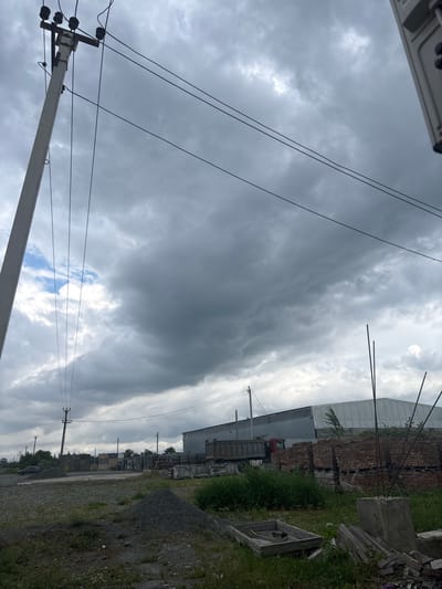 This is a tilted, outdoor photograph taken in Gizel, Russia. The scene depicts an industrial or construction area under a cloudy sky. In the foreground, a concrete utility pole with multiple insulators and wires is visible, angled diagonally across the frame. More power lines crisscross the sky. The middle ground features a large, light-colored industrial building with a corrugated roof, alongside a stack of red bricks and a truck. A narrow, unpaved road runs alongside a grassy verge and a pile of gravel. The sky is filled with dramatic, dark grey storm clouds, with a few patches of lighter sky and what appear to be faint rays of sunlight breaking through in the distance. The overall atmosphere is somber and overcast, suggesting a pre-storm or post-storm condition. No people are visible in the image. The time of day is not explicitly indicated but the lighting suggests it could be daytime.