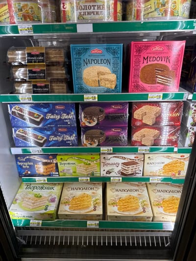 The image displays the interior of a refrigerated display case filled with various cakes and desserts. The items are arranged on multiple shelves. In the top shelf, several cylindrical packages with gold and red labeling are visible. The second shelf from the top holds packaged cakes, some labeled "Honey cake" and others with blue and pink boxes featuring descriptions like "NAPOLEON" and "MEDOVIK." The third shelf contains more packaged cakes, including "Fairy Tale cake" and "NIGHT." The fourth shelf presents "PERSIDSKAYA NOCH," "TVOROZHNIK," and "NAPOLEON" cakes in boxes with colorful illustrations. The bottom shelf shows more "TVOROZHNIK" and "NAPOLEON" cakes. The overall environment appears to be a retail setting, likely a grocery store or bakery, given the organized display of food products in a refrigerated unit. There are no people visible in the image. The specific location of Naperville, United States of America, is not determinable from the visual information presented in the image.