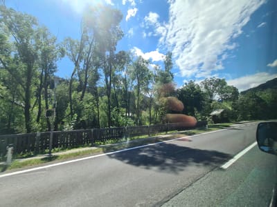 The image captures a sunlit scene from inside a vehicle, likely moving along a paved road in or near Dürnstein in der Steiermark, Austria. The road surface is asphalt, with a clear white line marking the right edge. To the left of the road is a grassy shoulder, followed by a wooden fence composed of vertical posts. Beyond the fence, a dense area of green trees rises, some of which appear tall and slender. In the background, more trees and vegetation cover a gentle hillside. The sky is a bright blue, with scattered white cumulus clouds and a strong sun glare, suggesting midday. On the far right of the image, a portion of the vehicle's side mirror is visible. In the distance, a small, rustic wooden structure with a sloped roof stands amidst the trees. The overall impression is a rural or semi-rural landscape on a clear day.