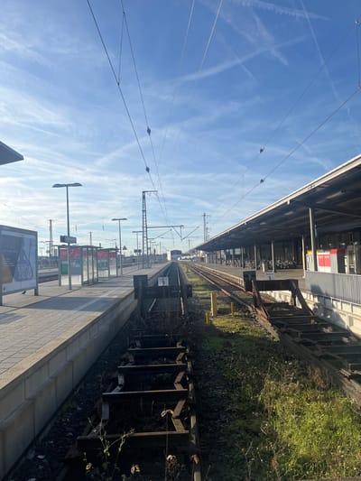 The image captures a static scene at a train station platform and tracks under a clear, bright blue sky in Schweinfurt, Germany.

The expansive sky, dominating the upper left portion, is adorned with several distinct white contrails or wispy cirrus clouds. Numerous intricate overhead catenary lines, supported by metallic poles and structures, crisscross the sky and extend along the railway.

A long, tiled train platform stretches along the lower left side of the frame, receding into the distance. On the platform, several modern streetlights with horizontal fixtures are visible, alongside multiple information boards. One board prominently features the German text "WINTER ZAUBER" (Winter Magic), while others display platform or track indicators such as "C 0 G," "1," and "2."

Adjacent to the platform, multiple sets of railway tracks, complete with ballast and wooden sleepers, extend into the horizon. Along the right side of the tracks, a strip of green vegetation and overgrown bushes adds a natural element to the industrial setting. A section of a station building or an overhead shelter, featuring a grey roof and hints of red signage, is partially visible above the tracks on the right.

The scene is well-lit by bright daylight, indicating a sunny day. No people or trains are present, contributing to a quiet and peaceful atmosphere at the station.