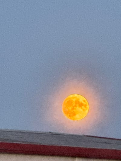 A full moon, appearing orange and luminous, is visible in a pale blue sky. The moon is partially obscured by a reddish-brown horizontal structure in the foreground, which may be a rooftop or a building edge. The sky exhibits a subtle textured pattern. The image appears to be a photograph taken in the city of Cherven, Belarus.