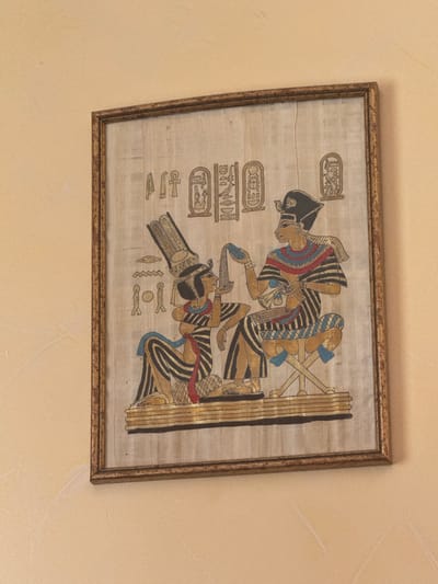 A framed, rectangular artwork depicting an ancient Egyptian scene on papyrus is mounted on a light yellowish-beige, textured wall. The artwork features two figures in profile, rendered in a palette of black, gold, red, and blue. On the left, a female figure with a tall feathered headdress and striped gown kneels, holding a blue strip of cloth or sash near her face. On the right, a male figure wearing a pharaonic crown and striped gown sits on a cross-legged stool, holding a small blue object in his left hand. Hieroglyphic cartouches and individual symbols are visible in the upper background of the artwork. The frame is ornate with a golden, slightly distressed finish. The wall exhibits subtle stucco-like texture and a faint diagonal scuff mark is present on the lower left portion. This scene is located in Aragats, Armenia.