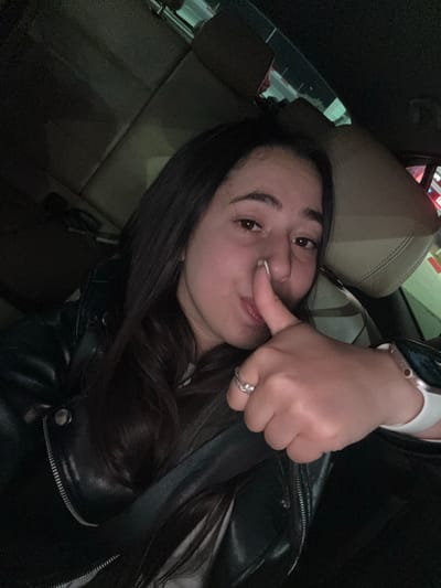 A selfie of a woman inside a car. She is wearing a black leather jacket and a watch on her left wrist, and a ring on her left ring finger. She is touching her nose with her left index finger. The car interior is visible, including a beige seat. The location in the city of Vodenichane, Bulgaria cannot be determined from the image.