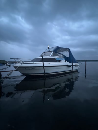A white motorboat named "King Detti" is docked on calm water under a dark, cloudy sky. The boat has a blue canvas cover over its upper deck. The surrounding environment appears to be a lake or river, with trees visible in the distance on the horizon. The scene suggests an overcast or stormy weather condition, likely in the late afternoon or early evening, given the low light. The boat is tethered to wooden posts, indicating it is moored. The water reflects the boat and the sky, creating a mirrored image. The location is Schwielowsee, Germany, a municipality known for its lake.