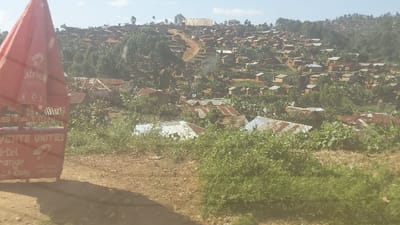 The image shows a view of a hillside in the city of Rutshuru, Democratic Republic of Congo, with coordinates -0.696 latitude and 29.172 longitude. The hillside is densely populated with a cluster of small houses, predominantly with corrugated metal roofs, and a few trees in between. In the foreground, there is a red sign that reads "Vente Unites Airtel Orange CD.COM". The sign is partially obscured by a red umbrella. The image appears to have been taken from a road, possibly a dirt road, with a shadow cast by the sign in the foreground. The perspective of the image suggests that the viewpoint is positioned higher than the surrounding structures, possibly overlooking the hillside from a slightly higher elevation.