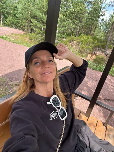 A blonde woman with fair skin and blue eyes wears a black baseball cap and a black sweatshirt. She has large hoop earrings and white sunglasses with dark lenses are hooked onto the collar of her sweatshirt. The woman is sitting on a wooden bench outdoors, with a wooded area and a reddish gravel path visible in the background. The woman is looking towards the camera with a slight smile and her right hand is resting on the brim of her cap. The location is identified as the city of Peski, Russia.