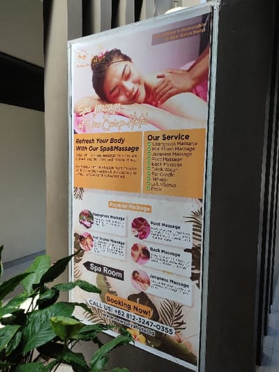A poster advertising spa services is attached to an exterior wall of a building in Gambiran, Indonesia at latitude -8.434 and longitude 114.18. The poster, which is made of white paper, is framed with a black metal frame and features a photograph of a woman in the upper left corner lying face up on a massage table receiving a massage. The woman has long black hair and is wearing a white top. The poster is advertising spa services at the "New Surya Hotel". The services are listed on the poster, including a range of massages such as deep tissue massage, hot stone massage, Javanese massage, foot massage, back massage, and full body massage. The poster also advertises a "Popular Package". The poster includes a green check mark next to each service. The poster also features a phone number and website for booking appointments. The poster is partially obscured by green foliage in the foreground. The wall is made of brown and beige tiles and has a texture similar to stone. The wall is partially obscured by a gray-brown wall in the background.