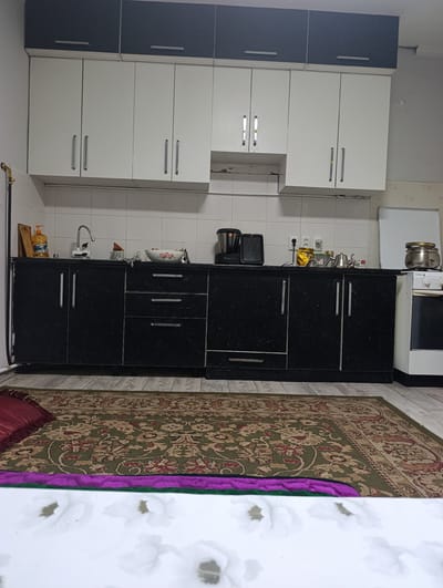 The image depicts a kitchen interior, likely in Lebedinovka, Kyrgyzstan. The kitchen features dark-patterned lower cabinets and white upper cabinets with dark gray accents. A white tiled backsplash runs along the wall behind the countertop. Various kitchen appliances and items are visible on the countertop, including a faucet, a bottle of dish soap, a blender, a coffee maker, a teapot, and a pot on a white stove. A patterned rug covers the floor in front of the cabinets. The overall scene is of a functional, modestly equipped kitchen.