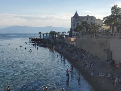 This is a photograph of a busy beach scene in Loutra Edipsou, Greece, likely during the late afternoon given the long shadows and warm, golden light. The water is calm and blue, with many people swimming and wading near the shore. Some individuals are further out in the sea, enjoying the water.
The beach itself is a mix of sand and rocky areas. Along the shore, numerous people are sunbathing, relaxing on towels, and walking near the water's edge. A distinctive, multi-story building with a conical roof and white facade dominates the background, suggesting a hotel or a historic structure. Palm trees and other lush greenery are scattered along the beachfront and around the building, adding to the tropical atmosphere.
A wooden pier extends into the sea from the left side of the image, with people visible on it. A pathway or promenade runs along the elevated area behind the beach, bordered by a white railing, where more people are strolling and observing the scene. A lifeguard stand or similar structure is visible on the right side of the beach. The sky is a clear blue with a few wispy clouds, and distant mountains are visible on the horizon. The overall mood is one of relaxation and enjoyment, with a clear sense of a popular tourist destination. There is no visible text in the image.