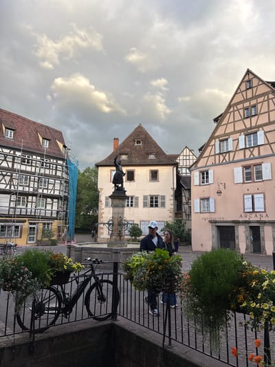 The image depicts a scene in the city of Colmar, France, likely in the historic old town. In the foreground, a black bicycle is parked next to a black metal railing adorned with hanging flower baskets overflowing with green foliage and colorful flowers. Behind the bicycle, a stone fountain with a statue of a person is visible, surrounded by cobblestone pavement.

Several timber-framed buildings with red-tiled roofs line the square. On the left, a building is covered in scaffolding and blue netting, indicating ongoing renovation. To the right of the fountain, a pale pink building features numerous windows with white shutters and signage reading "ANTIQUITES ARCANA."

In the mid-ground, two people, a man wearing a white cap and dark jacket, and a woman wearing glasses and a dark top, are walking away from the viewer. They are on the cobblestone area, in front of the buildings. The sky above is partly cloudy with a dramatic, overcast appearance, suggesting a late afternoon or evening setting. The architecture and the overall atmosphere point to a picturesque European town square.