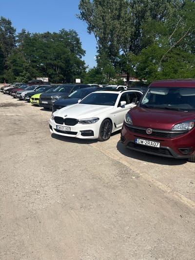 In the city of Kobyla Góra, Poland, on a clear, sunny day, a line of cars is parked on a gravel surface. The foreground features the front of a dark red Fiat van, displaying a license plate that reads "DW 2RA07". To its left, a white BMW station wagon is parked, its front bumper and license plate visible. The license plate on the BMW shows "DW 6Y J42". Behind the BMW, a row of other vehicles is visible, including a dark gray car with a lime green accent and other dark-colored cars. In the background, a line of green trees stands tall against a bright blue sky. The overall scene depicts a parking area in a semi-rural or suburban setting.