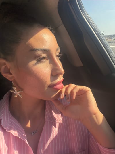 This media file features a close-up, sideways portrait of a woman's face and upper body, bathed in warm sunlight. She has her eyes closed and her lips are slightly parted, with one hand brought up to her mouth in a suggestive pose. Her face shows signs of natural skin texture with some visible pores and a subtle sheen of sweat or oil, indicating a warm environment. She is wearing a pink and white striped shirt, and a delicate silver necklace with the name "Yvone" is visible on her collarbone. A prominent detail is her earring, a gold starfish adorned with small white pearls.

The scene appears to be set inside a vehicle, suggested by the dark, curved frame and window visible in the upper left portion of the image, which resembles the interior of a car or van. The bright light suggests it is daytime, possibly late morning or afternoon, and the quality of the light hints at clear weather. The overall mood is relaxed and sensual, with the soft lighting and the woman's posture contributing to this impression. The image is oriented sideways. The location context provided is Aradippou, Cyprus.