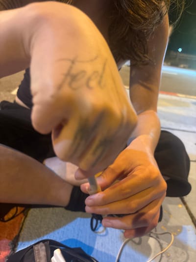 The scene depicts an individual seated outdoors, likely on a sidewalk or paved area, at night in Thap Sakae, Thailand. The person is partially visible, wearing a dark top and dark pants, with long, brown hair visible on the right. Both hands are engaged in manipulating a slender, light-colored cylindrical object. The left hand, closer to the viewer, displays a tattoo reading "Feel" on its back and blurred dark markings on the fingers, gripping one end of the object. The right hand holds the other end, possibly inserting it into a wider, lighter-colored sleeve or tube. A black bag or backpack is visible on the ground to the lower left, containing a white rectangular object and accompanied by tangled white and black cables. The ground features blue and yellow painted markings. The background is out of focus, showing a dark street illuminated by distant artificial lights, casting an amber glow on the immediate foreground.