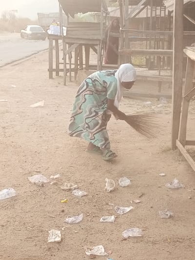 The image depicts an outdoor scene in a dusty, unpaved area, likely a roadside market or vendor space in Ebiya, Nigeria. In the foreground, a woman is actively sweeping the ground with a broom made of natural fibers. She wears a white head covering (hijab or scarf), a long, patterned dress in shades of teal/mint green and dark brown/black, and green sandals. The ground around her is heavily littered with numerous crumpled plastic bags and other small pieces of refuse.
Behind the sweeping woman, several rustic wooden structures, resembling market stalls or shelters, are visible. Inside one of these structures, a second woman, dressed in a dark red garment, is partially visible. Further in the background, a hazy road can be seen, with a dark-colored sedan car on it. The overall atmosphere is characterized by a significant amount of haze or dust, which blurs the background elements. The scene portrays a routine cleaning activity in a functional, open-air setting. No text is visible in the image.