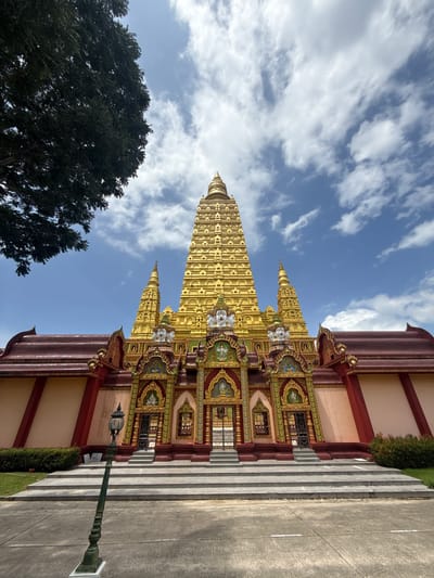 The image shows the exterior of a Buddhist temple in Na Nuea, Thailand. The temple features a prominent golden stupa with intricate architectural details and multiple tiers. The lower section of the temple is adorned with colorful decorations and statues. A wide set of stone stairs leads to the entrance. In the foreground, a dark green lamppost stands on a concrete surface. Lush green bushes and a large tree with dark foliage are visible on the left side of the temple. The sky is blue with scattered white clouds. The scene is captured from a low angle, emphasizing the grandeur of the temple.