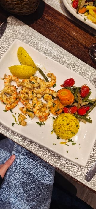 A rectangular white plate displays a vibrant meal. A mound of yellow rice sits near a cluster of green beans, cherry tomatoes, and orange carrots. A generous portion of seasoned shrimp, topped with green herbs, rests beside two lemon wedges. The plate is placed on a textured gray placemat, with the edge of a wooden table visible. A glimpse of someone's blue jeans and hand adds a personal touch to the scene.