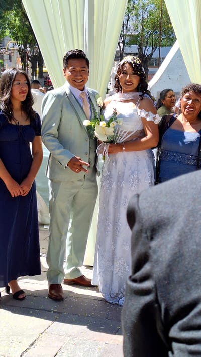 Captured in Cerro Colorado, Peru, the image portrays a wedding celebration outdoors. A newlywed couple stands at the center. The bride wears a white floral lace gown and holds a bouquet of white calla lilies. The groom, in a light green suit, flashes a peace sign. A woman in a blue dress stands below the groom and beside a woman in a blue dress stands above the bride. Sprinkled confetti dots their heads and shoulders. They stand under a light yellow canopy. The foreground shows the side of someone in grey. Trees are visible in the background.