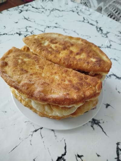Two fried pastries, likely chebureki, are stacked on a white plate, which sits on a white tablecloth with a black marble-like pattern. The pastries are golden brown with slightly bubbly surfaces, indicating they have been fried. The lower pastry is partially visible, revealing a lighter colored filling or dough layer. The background is out of focus, but the tablecloth and hints of a colored surface suggest an indoor setting. There are no people or specific landmarks visible that would indicate a location in Ceadîr-Lunga, Moldova.