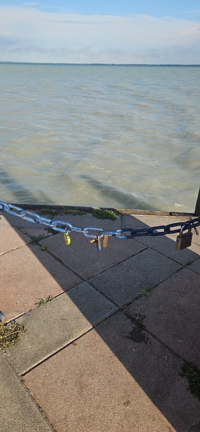 A chain secured with multiple padlocks stretches horizontally across the foreground of the image. The chain is attached to a wooden post on the right and extends out of frame to the left. The chain is linked to a pier or walkway, made of interlocking pavers in shades of pink, gray, and tan. Small patches of green grass grow between the pavers. In the background, a large body of water, likely Lake Balaton, stretches to the horizon under a pale blue sky with wispy clouds. The water is choppy, with visible ripples and waves. A thin line of trees or vegetation is visible on the distant shore. The scene is captured from a low angle, looking out over the water. The overall impression is one of a serene, outdoor location, possibly a public access point to the lake.