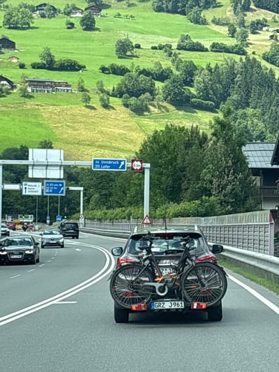 This is a vertical, eye-level shot of a road in Zell am See, Austria, on a bright, possibly sunny day. The road curves to the right, with a white guardrail on the left and a grassy, treed hillside on the right.

The main focus of the image is a dark-colored SUV or van stopped on the right side of the road. The rear of the vehicle is prominent, and it is carrying two bicycles on a rack. The license plate is visible and reads "GRZ 3961".

Several road signs are visible. One blue sign points to "Innsbruck" and "Lofer", with a height restriction of "4.0m". Another blue sign indicates directions to "Zell am See", "Lofer", and "Kaprun Schützdorf". A smaller circular sign with a red border and a "4.0m" marker is also present.

In the background, the green, rolling hills are dotted with trees and some buildings, suggesting a rural or semi-rural Austrian landscape. Several other vehicles, including a black car and a black taxi, are also visible on the road, indicating traffic. The overall impression is a scenic drive in the Austrian Alps.