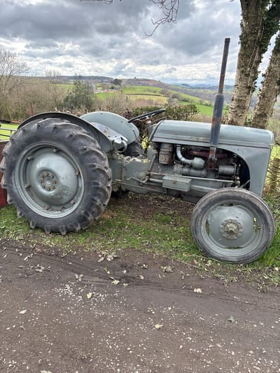 A gray, vintage tractor is parked on a grassy verge adjacent to a dirt road in Stiperstones, United Kingdom. The tractor is viewed from the side, with its large rear tire prominent and a smaller front tire also visible. The engine and exhaust pipe are detailed, exhibiting signs of age and rust. The background shows a landscape of green fields and distant hills under a cloudy sky. Some trees and hedges border the field, adding depth to the scene.
