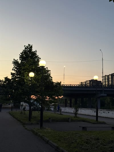 The image depicts a street scene in Kamysh-Gadzhiaul, Russia, likely during dusk or early morning given the soft, warm light illuminating the sky. The foreground features a paved pathway bordered by a grassy embankment and trees with dense foliage. Several modern streetlights with spherical lamps are visible, two of which are brightly lit, casting a warm glow. In the mid-ground, a large bridge spans across a body of water or a significant urban divide. The bridge is characterized by its dark, structural elements and a railing. Beyond the bridge, a multi-story building with numerous windows is silhouetted against the sky. The overall mood of the image is serene and quiet, with the artificial lights creating a sense of atmosphere. The presence of a person on a bicycle in the mid-ground suggests some activity, but the scene primarily conveys a sense of calm urban environment at the transition between day and night. There is no visible text in the image.