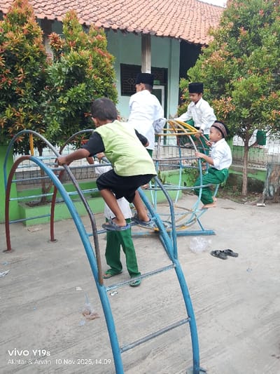 Four children play on playground equipment. One, wearing a green shirt and black shorts, climbs a blue ladder structure. Another, wearing a white shirt and green pants, stands at the base of the ladder. Two more children in white shirts and green pants are on a connected circular structure. Some children wear black peci hats. Discarded plastic wrappers and shoes lie on the concrete ground. Trees with red-tipped leaves and a building with a red tile roof form the background. The date "10 Nov 2025" is stamped on the lower left corner.