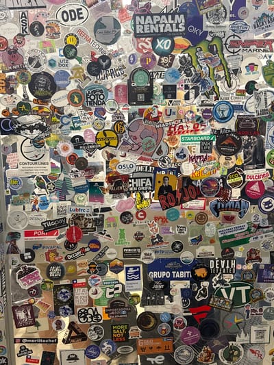 A chaotic collage of stickers densely covers a surface. The stickers are of varied shapes, sizes, and designs, including brand logos, travel destinations, slogans, and artwork. Overlapping and jumbled, the stickers create a visually busy and textured background, with a mix of colors and themes. Some discernible stickers feature destinations like Oslo and Lake St. George, brands like Napalm Rentals and Lubrtc, and slogans like "Come Follow Me" and "Let All That You Do Be Done In Love".