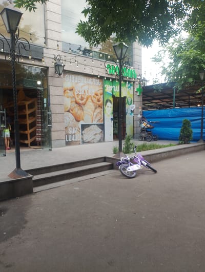 The image shows a street scene in the city of Nor Hachin, Armenia. In the foreground, a purple child's bicycle lies on its side on the pavement. Behind the bicycle, there is a set of concrete steps leading up to a building. The building appears to be a store, with large glass windows and a sign with text in Armenian. The store has shelves stocked with bread products visible through the windows. Further down the street, a stroller is parked next to a small evergreen tree. The background includes a blue tarp-covered structure, possibly a market stall or temporary roofing. Overhead, tree branches with green leaves hang over the scene, and decorative lights are strung along the building's facade. A street lamp with a decorative metal post is positioned to the left of the frame.