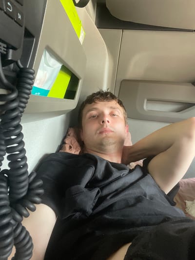 The image shows a young man lying down inside a truck cab. He is wearing a black t-shirt and appears to be resting. The interior of the truck is visible, including a coiled black cord, a tissue dispenser with a green box, and what appear to be storage compartments. The lighting suggests it is daytime. The location is not specified as Brachterbeek, Netherlands in the image.