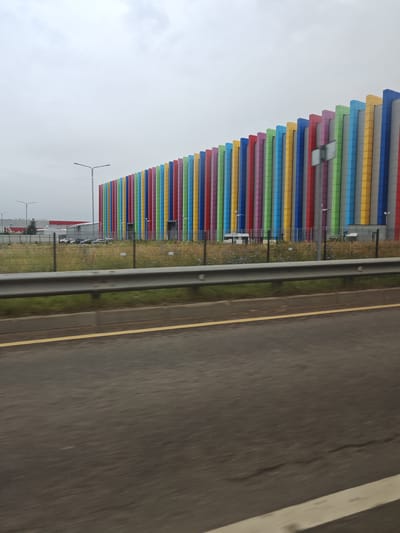 The image shows the exterior of a large, modern building with a distinctive facade composed of vertical, brightly colored panels in shades of blue, green, yellow, red, and pink. The building appears to be an industrial or commercial facility, as indicated by the presence of large doors and the surrounding infrastructure. In the foreground, a road with a yellow line is visible, suggesting the photo was taken from a vehicle in motion. A metal guardrail separates the road from a grassy area behind which the building stands. Several cars are parked in front of a smaller, adjacent building to the left of the colorful structure. Streetlights are visible in the middle ground. The sky is overcast, and the overall impression is that of a functional, somewhat utilitarian urban or suburban landscape. The building's vibrant facade is its most striking feature.