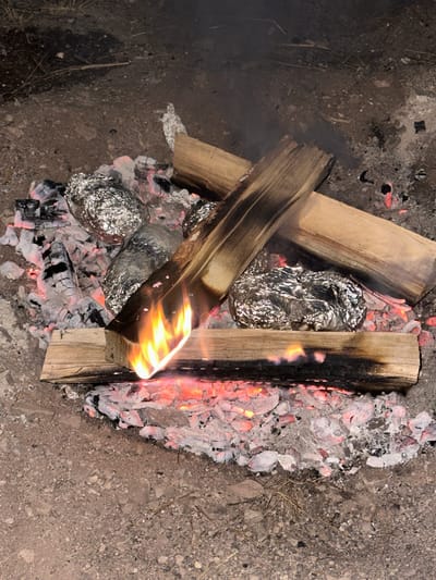 A close-up, high-angle shot captures a campfire with several potatoes wrapped in aluminum foil being cooked in the glowing embers and ashes. The fire is actively burning with visible flames licking up the side of a piece of wood. Several logs are arranged around the potatoes. The surrounding ground is composed of dirt and small pebbles. There are no people or animals visible in the frame. The scene appears to be outdoors.
