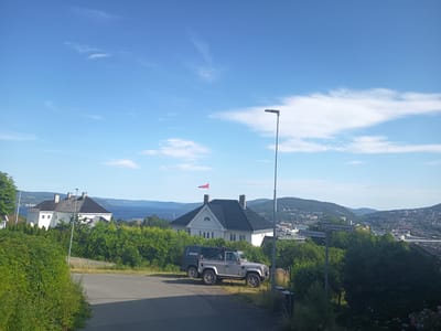 The image depicts a scenic view of Drammen, Norway on a clear, sunny day. The foreground is dominated by a paved road and lush green bushes. Two vehicles, a dark colored truck with "ROCKTON" written on its side and a silver off-road vehicle, are parked on the side of the road. Behind the vehicles, a white house with a dark roof and dormer windows is visible. A Norwegian flag flies from the roof of the house. In the background, a body of water, likely a fjord, is visible, with rolling hills and the city of Drammen spread out on its slopes. The city appears to be built into the hillside, with numerous buildings and infrastructure, including a bridge. Two street signs are visible: "Frydenberggata" and "Strøtvetveien," with "Oppfartveien" also visible above Frydenberggata. The sky is a vibrant blue with scattered white clouds, suggesting it is daytime, likely late morning or early afternoon. The overall impression is one of a peaceful, picturesque residential area with a stunning natural backdrop. There is no visible activity or people in the image, creating a serene and tranquil atmosphere. The time of day is indicated by the bright sunlight and clear sky, typical of a summer day in Norway.