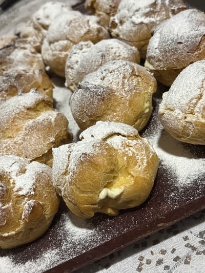 This image shows a close-up view of several cream puffs dusted with powdered sugar. The cream puffs are golden brown and appear to be freshly baked. Some of them have a visible white cream filling peeking out. The powdered sugar is scattered unevenly across the surface of the cream puffs and on the dark brown tray they are resting on. The background is out of focus but appears to be a light-colored, textured surface, possibly a tablecloth. There are no people, animals, or specific landmarks of Sheerteba, Georgia visible in the image. The scene is entirely focused on the arrangement of these pastries.