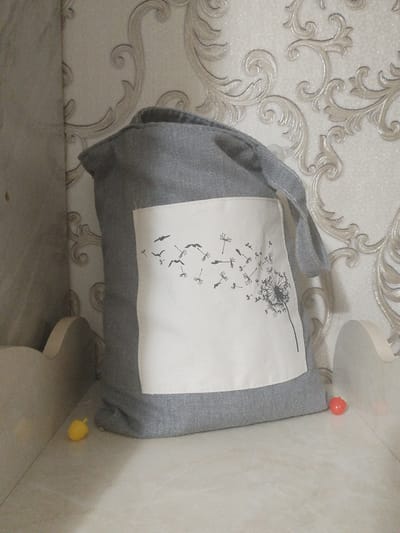A grey fabric bag with a white rectangular patch on the front featuring a black dandelion silhouette design rests on a shelf against a cream-colored wallpaper with an ornate swirling pattern. Two small, brightly colored objects, one yellow and one red, are placed near the base of the bag on the shelf. The bag has two grey fabric straps. The environment appears to be an indoor setting, possibly a room in a home. There are no people visible in the image. The scene is static and the lighting is soft.