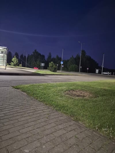 The image shows a street scene at night in Pabradė, Lithuania. In the foreground, a paved walkway made of grey bricks runs along the left side of the frame, adjacent to a strip of green grass. Beyond the grass is a paved road. In the background, several streetlights illuminate the area. On the left side of the road, there is a bus stop shelter with glass panels. Further down the road, a red car is parked. Across the road, more trees line the landscape, and several traffic signs and streetlights are visible. A silver car is parked near a bus stop shelter on the right side of the road. The sky is a deep blue, indicating it is nighttime, with a faint atmospheric phenomenon visible in the upper left portion of the sky.