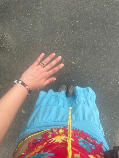 A first-person perspective shot shows a person standing on an asphalt surface, with their hand and arm extended outwards towards the left of the frame. The hand displays two rings on the index and middle fingers, and a bracelet made of multi-colored beads adorns the wrist. The person is wearing a bright blue skirt and a colorful top with a floral pattern. Gray sneakers are visible on their feet. The background is an out-of-focus gray asphalt surface. The location is the city of Sempigny, France.