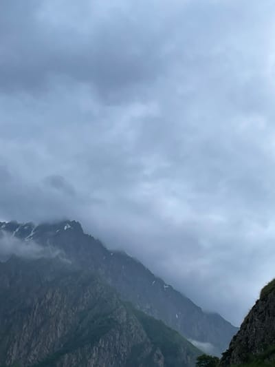 The image shows a dramatic mountain landscape shrouded in mist and clouds. The foreground features the rocky, steep slopes of a mountain on the right side of the frame, with some vegetation visible. The main focus is on a range of tall, jagged mountains that stretch across the middle ground, their peaks partially obscured by dense, gray clouds. Patches of snow are visible on the higher elevations of these mountains. The sky above is overcast, with heavy, layered clouds contributing to a somber and atmospheric mood. The overall impression is one of a remote, rugged, and somewhat moody natural environment, characteristic of the mountainous region of Tsdo, Georgia. There are no people or man-made structures visible in the image.