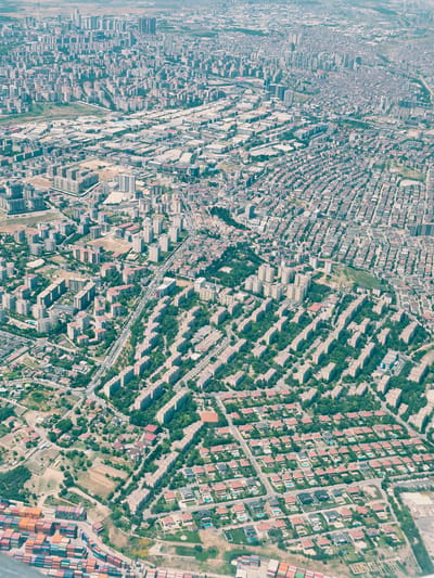 This is an aerial photograph of a dense urban landscape, identified as Beylikdüzü, Turkey. The image is dominated by an extensive grid of residential buildings, varying in size and architectural style. In the foreground, a collection of stacked shipping containers is visible, suggesting proximity to a port or industrial area. The midground features a uniform development of mid-rise apartment buildings, many with red-tiled roofs, arranged in rows with tree-lined streets and courtyards. Further into the background, the cityscape transitions to a mix of taller residential towers and commercial or industrial buildings, with a sprawling network of roads connecting them. The overall impression is one of high-density urban living with pockets of green spaces interspersed among the structures. There are no visible individuals or specific actions depicted in the photograph; it is a static overview of an urban environment. The lighting suggests daytime, possibly late morning or early afternoon, given the shadows cast by the buildings. The sky is hazy, which is common in urban areas.